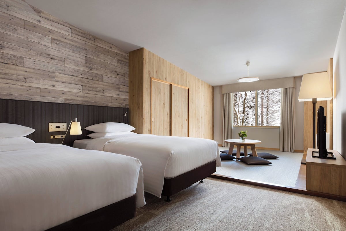 A hotel room with two beds, a wooden accent wall, a small table with a lamp, and a large window with a view of trees.