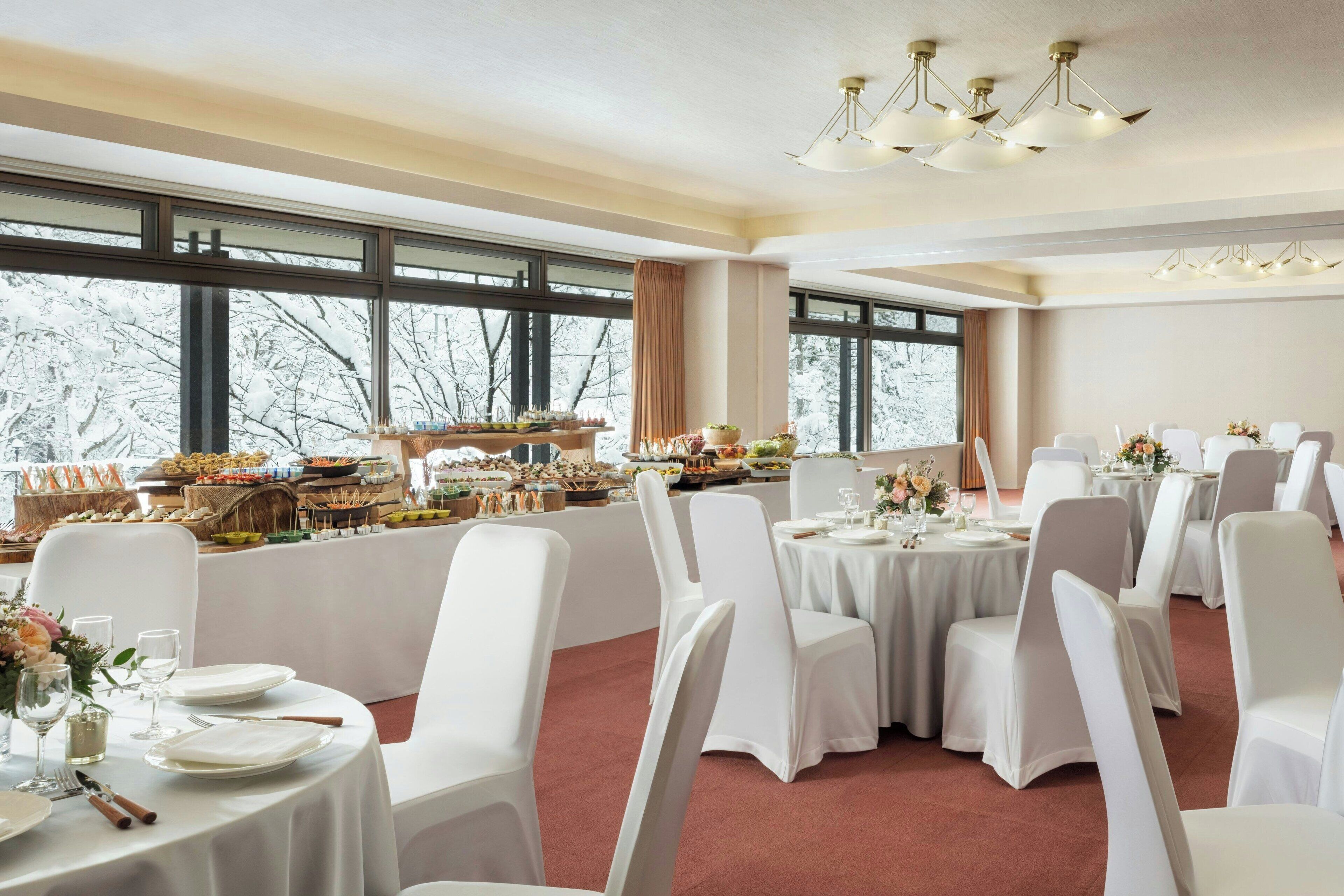 Un salón de banquetes con mesas redondas, sillas blancas y una zona de bufé.