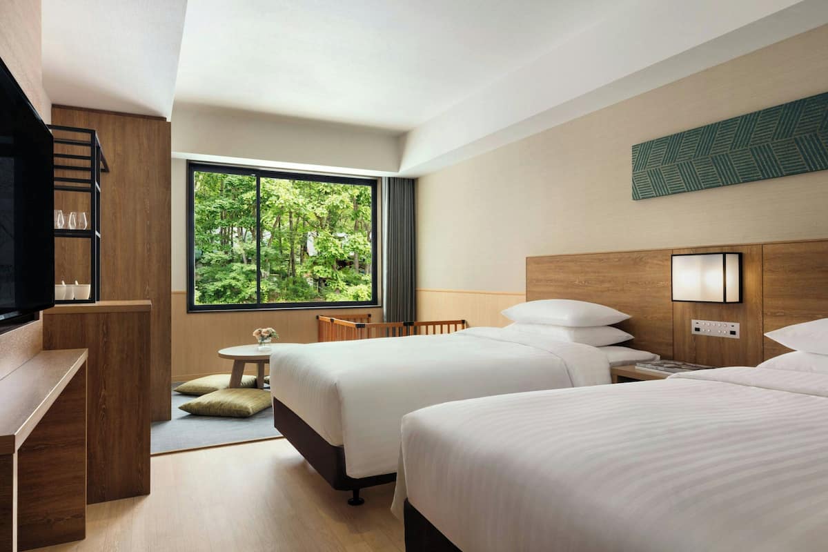A hotel room with two beds, a desk, a chair, a small table, and a large window offering a view of greenery.
