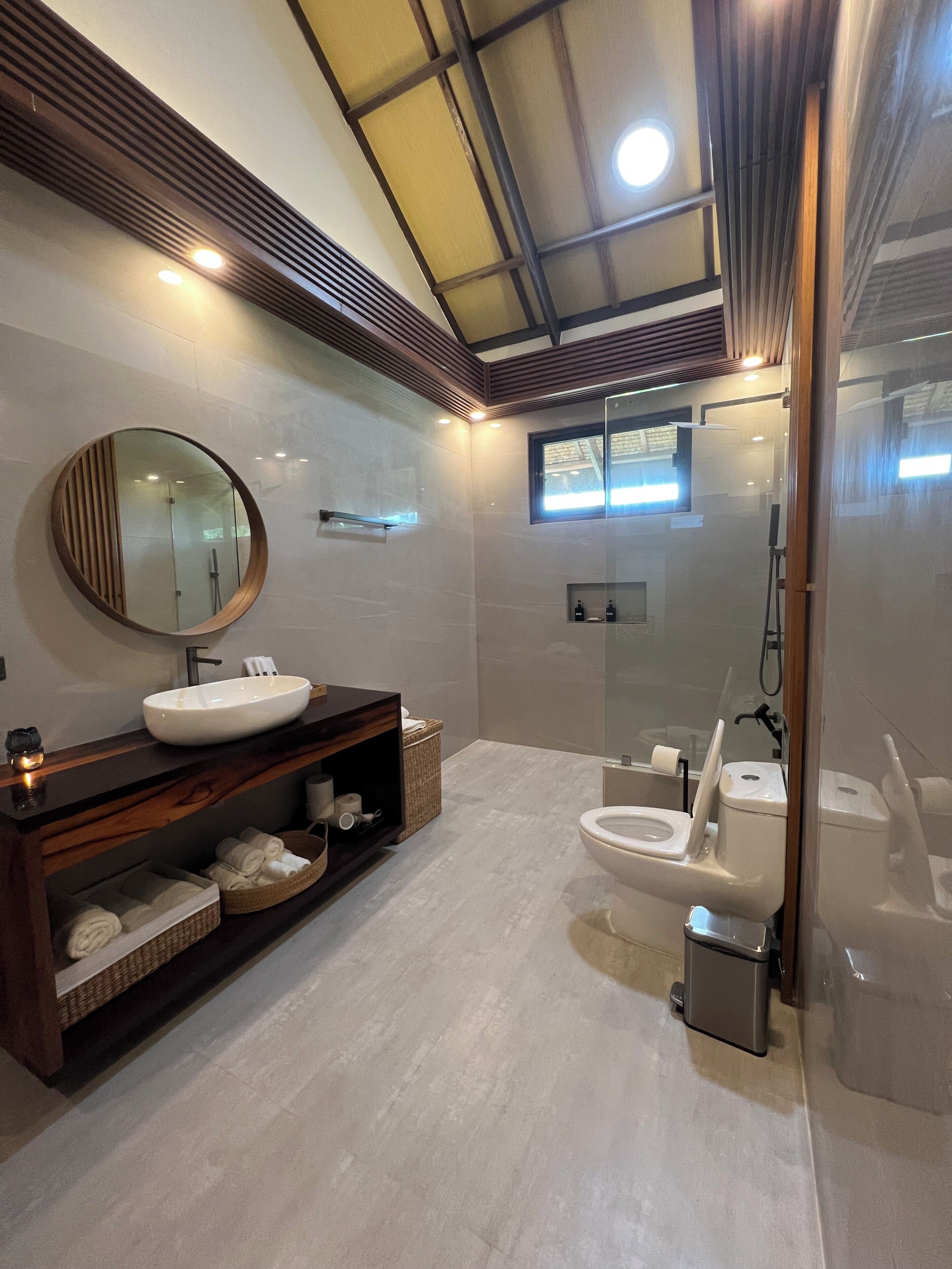 A modern bathroom with a round mirror, a vessel sink, a freestanding toilet, and a glass-enclosed shower area.