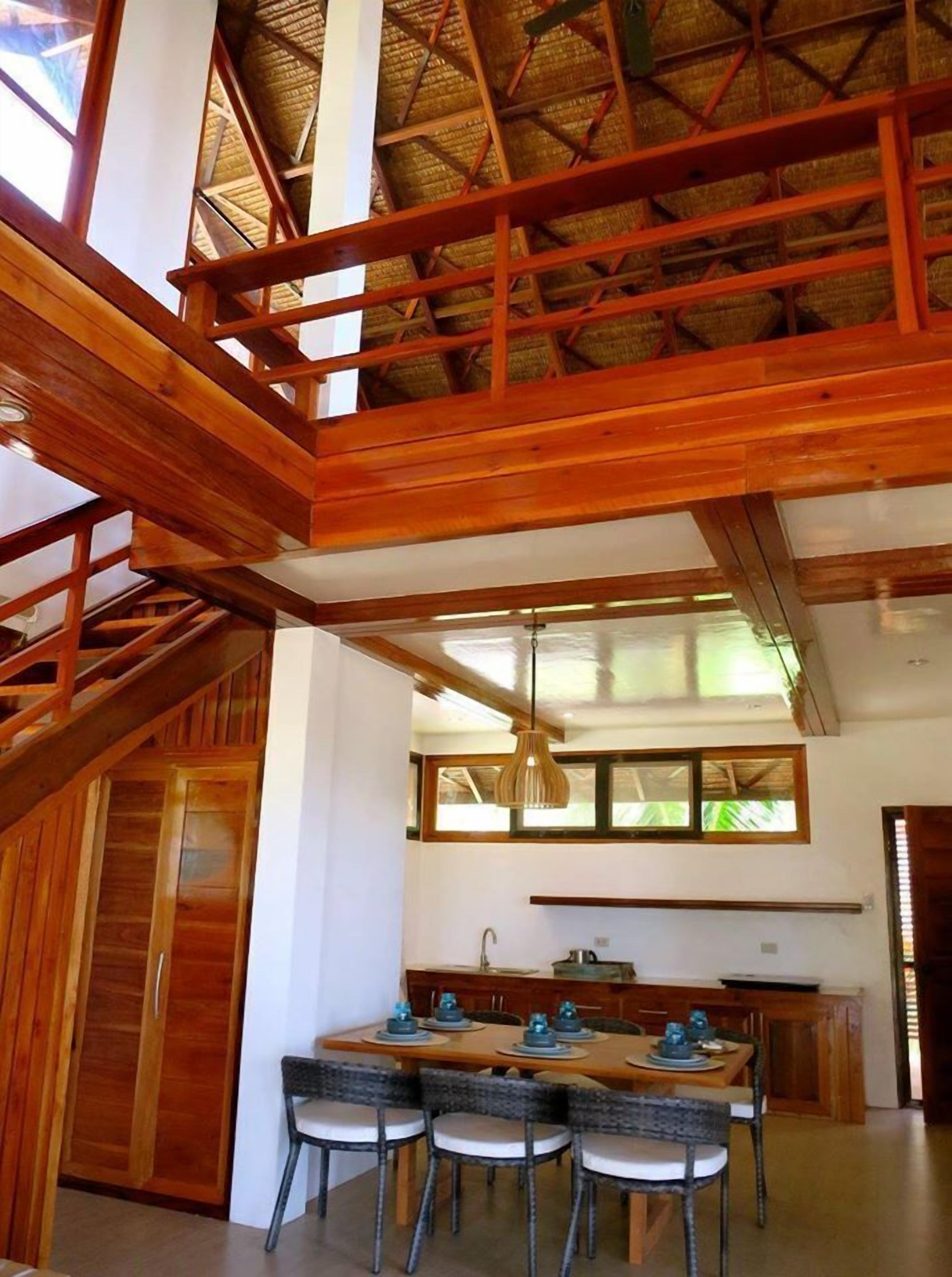 Ein geräumiger Essbereich mit hoher Decke und Holzbalken, eine Küche mit Fenster sowie eine Treppe, die in ein Obergeschoss führt.