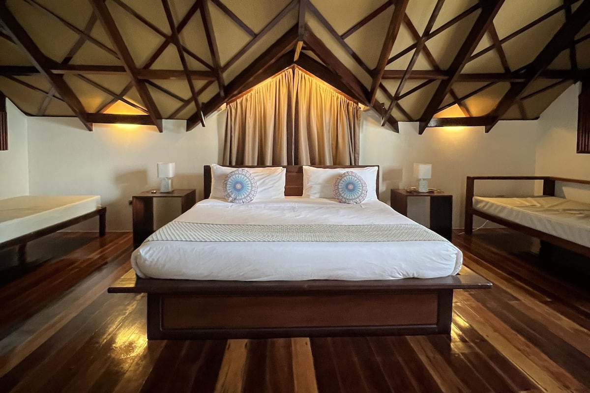 Un dormitorio con techo de madera, una cama grande con ropa de cama blanca, dos mesitas de noche con lámparas y suelo de madera.