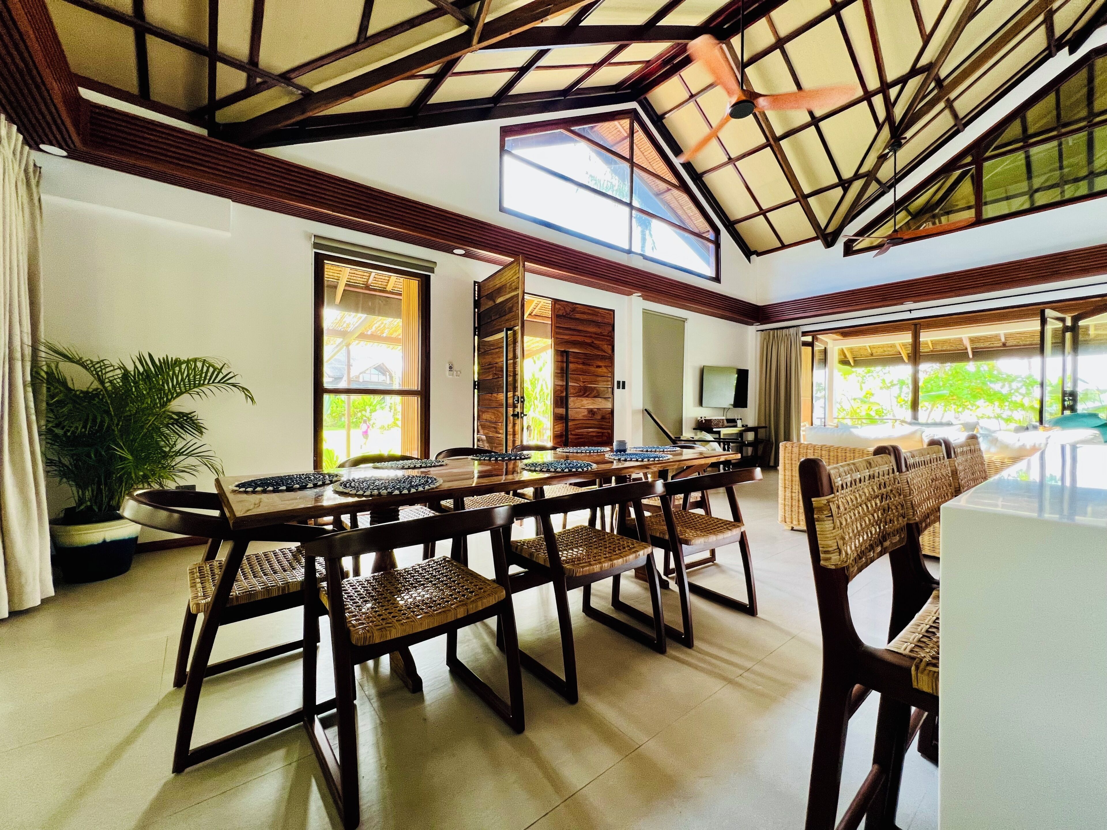 A spacious room with a high ceiling, large windows, and a dining area with a long table and wicker chairs.