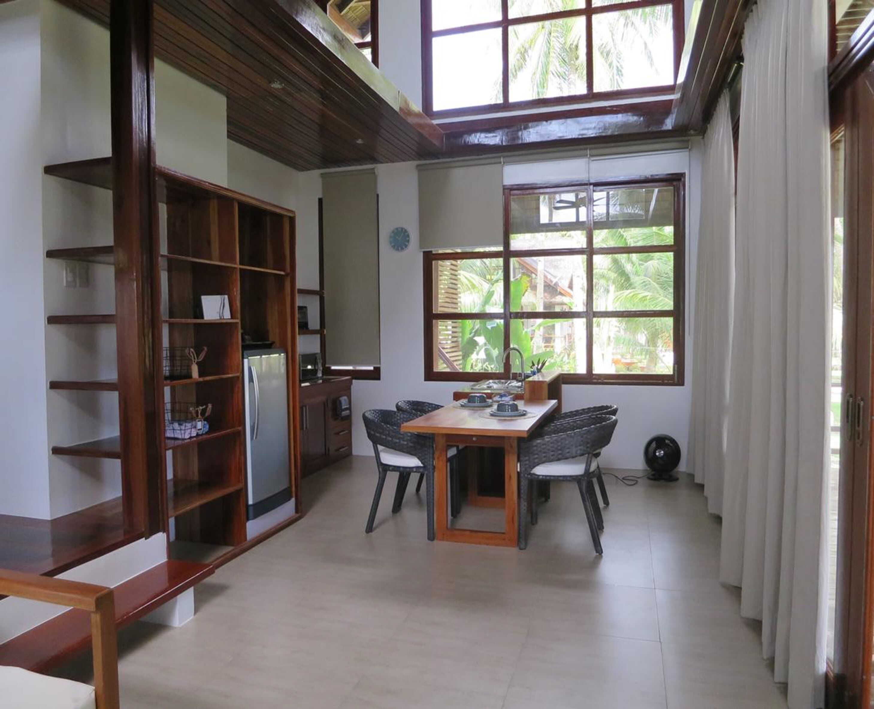A spacious room with a wooden dining table, two chairs, a bookshelf, and a large window with a view of greenery.