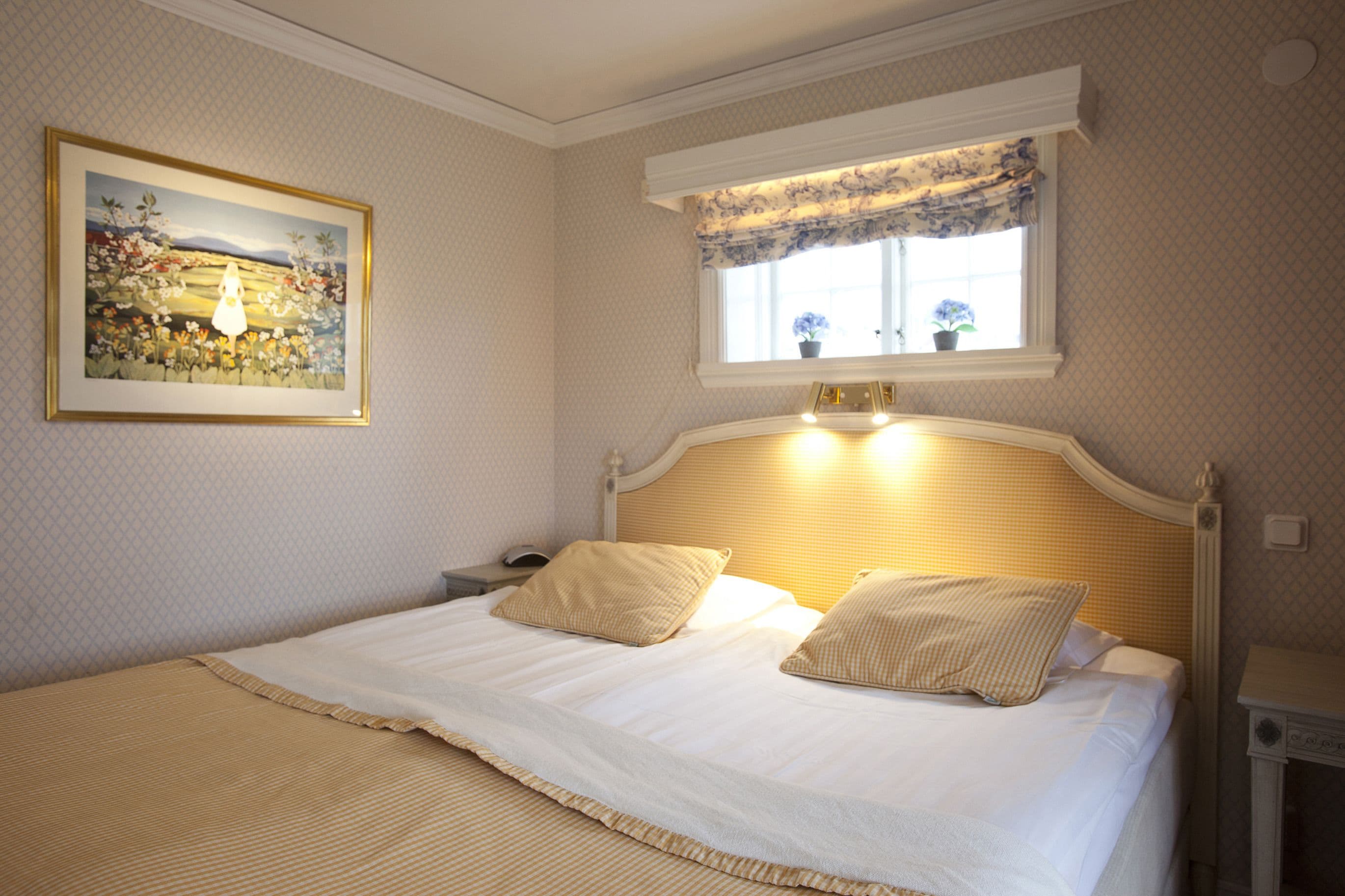 A neatly made bed with two pillows, a bedside table with a lamp, a framed picture on the wall, and a window with a floral curtain.
