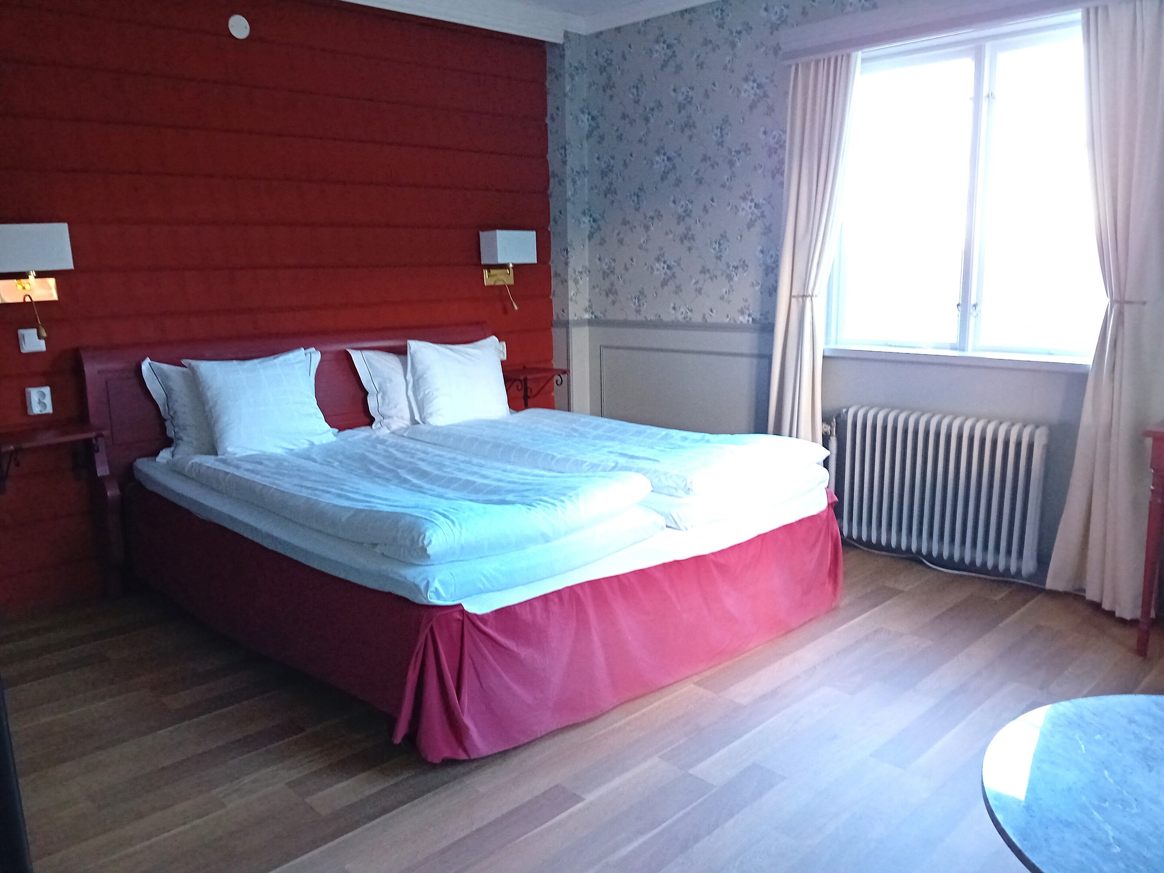Una cama en una habitación con cabecera roja, un radiador y una ventana con cortinas.