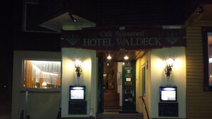 The exterior of Hotel Waldeck, a cafe restaurant with a sign above the entrance, two lit wall sconces, and a staircase leading to the door. - Hotel Waldeck (Todtmoos)