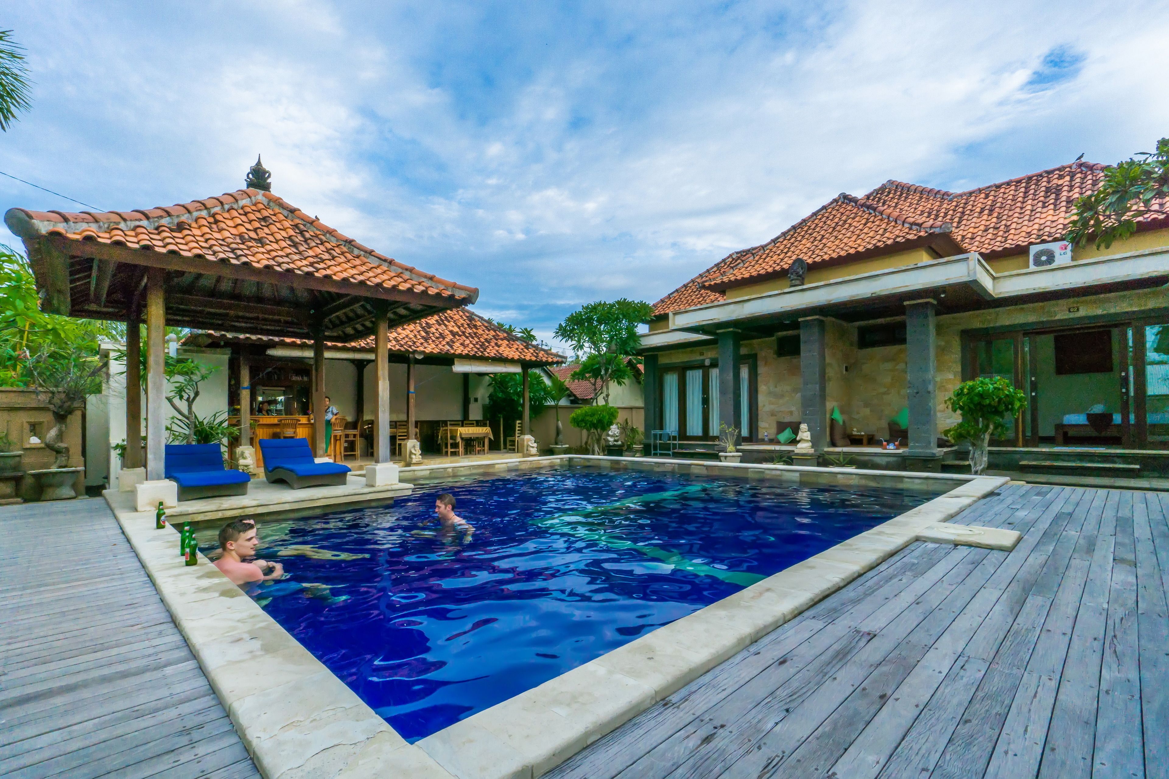 Une piscine avec une terrasse en bois, un espace salon couvert et un bâtiment doté d’un toit en tuiles.