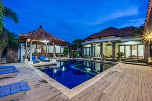 A swimming pool with lounge chairs, a covered seating area, and a building with a tiled roof.