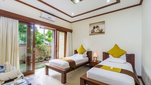 A hotel room with two beds, a small table, a desk, and a view of the garden through the sliding glass door. - Pondok Baruna Garden Rooms (Lembongan Island)