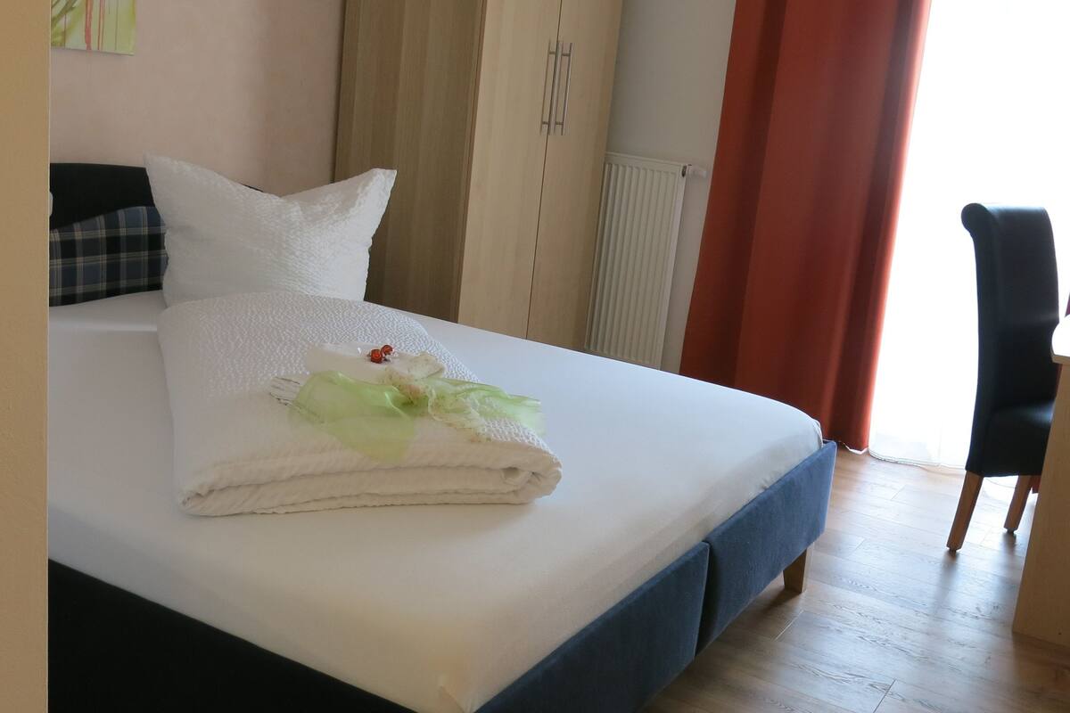 A neatly made bed with white linens and a folded towel, a plaid pillow, and a white pillow on a wooden floor with a radiator and curtains in the background.