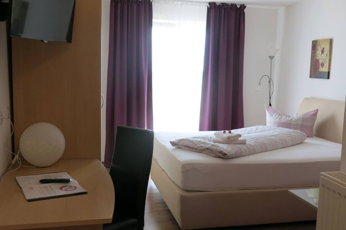 A hotel room with a bed, a desk with a chair, a television mounted on the wall, and purple curtains.