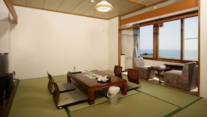 Ein traditioneller japanischer Raum mit Tatami-Böden, einem Holztisch, Stühlen, einem Fernseher und Meerblick.