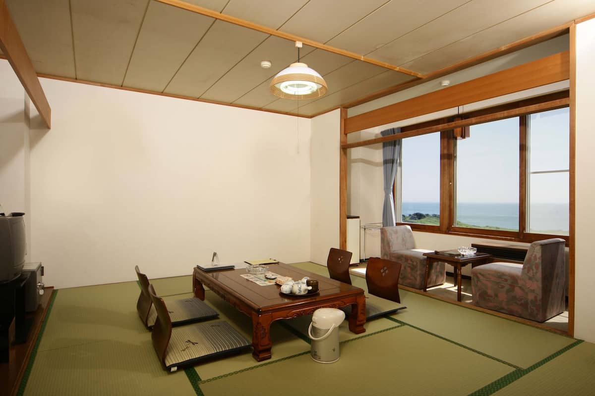 A traditional Japanese room with tatami flooring, a wooden table, chairs, a television, and a view of the sea.