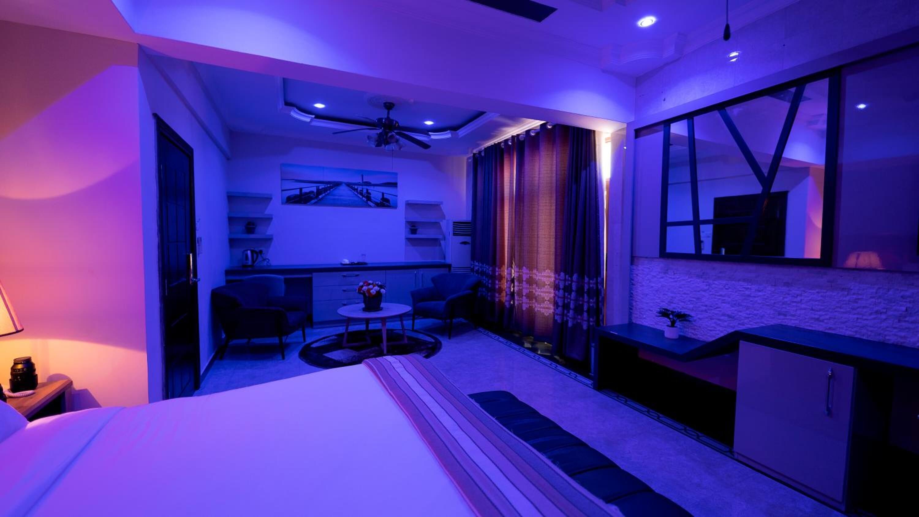 Una habitación de hotel moderna con cama, una zona de estar con mesa y sillas, una kitchenette pequeña y un ventanal con cortinas.