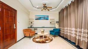 A living room with a wooden door, a ceiling fan, a round coffee table with a vase, two armchairs, and a view of a kitchen and dining area through a glass partition.