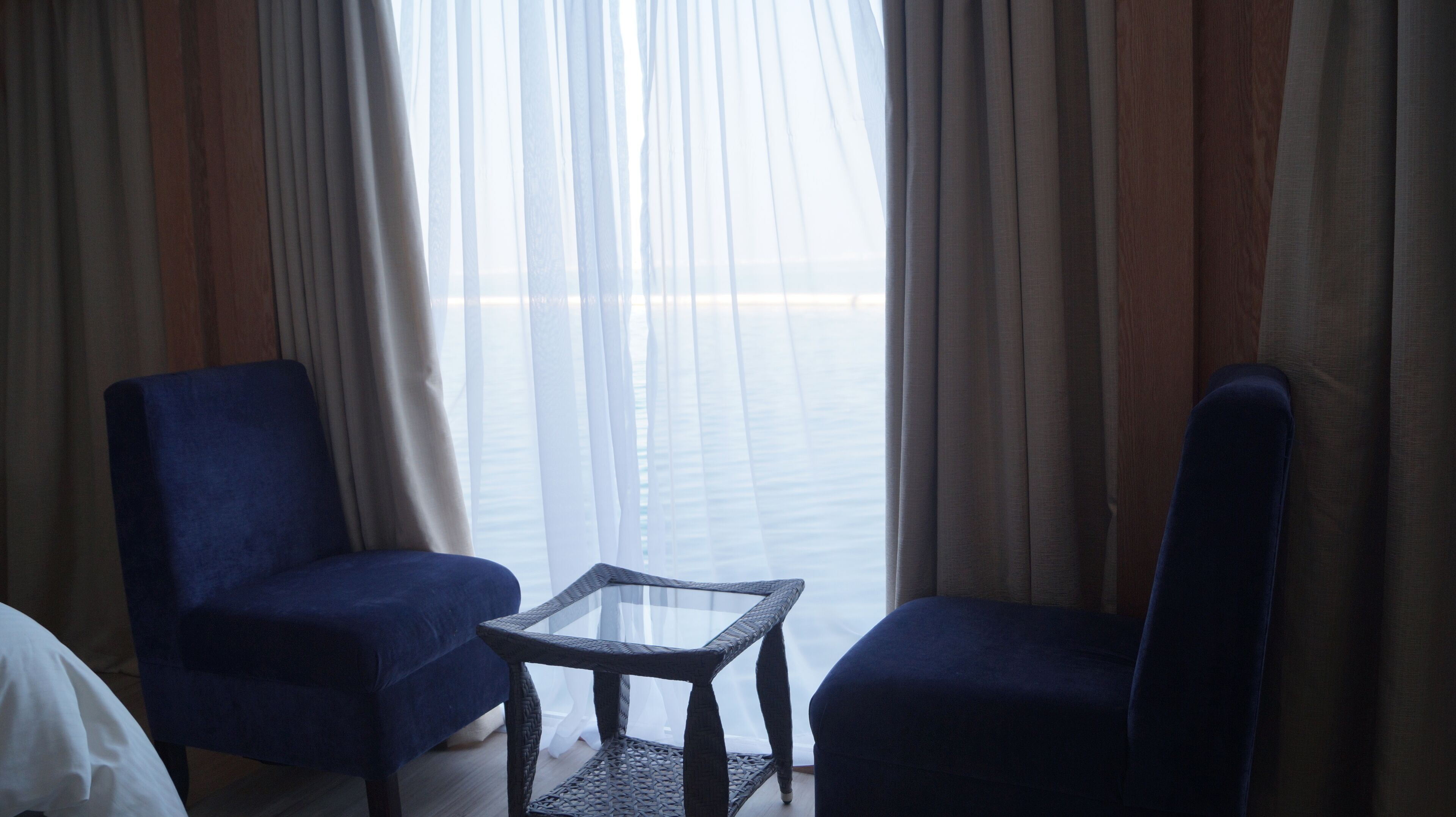 Une chambre d’hôtel avec deux fauteuils bleus, une petite table en verre et une vue à travers des rideaux transparents.