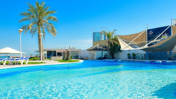 A swimming pool with lounge chairs, palm trees, and a building with a construction crane in the background. - C Hotel (Manama)