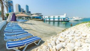 A beachside hotel with lounge chairs, a rocky shoreline, and boats in the water. - C Hotel (Manama)