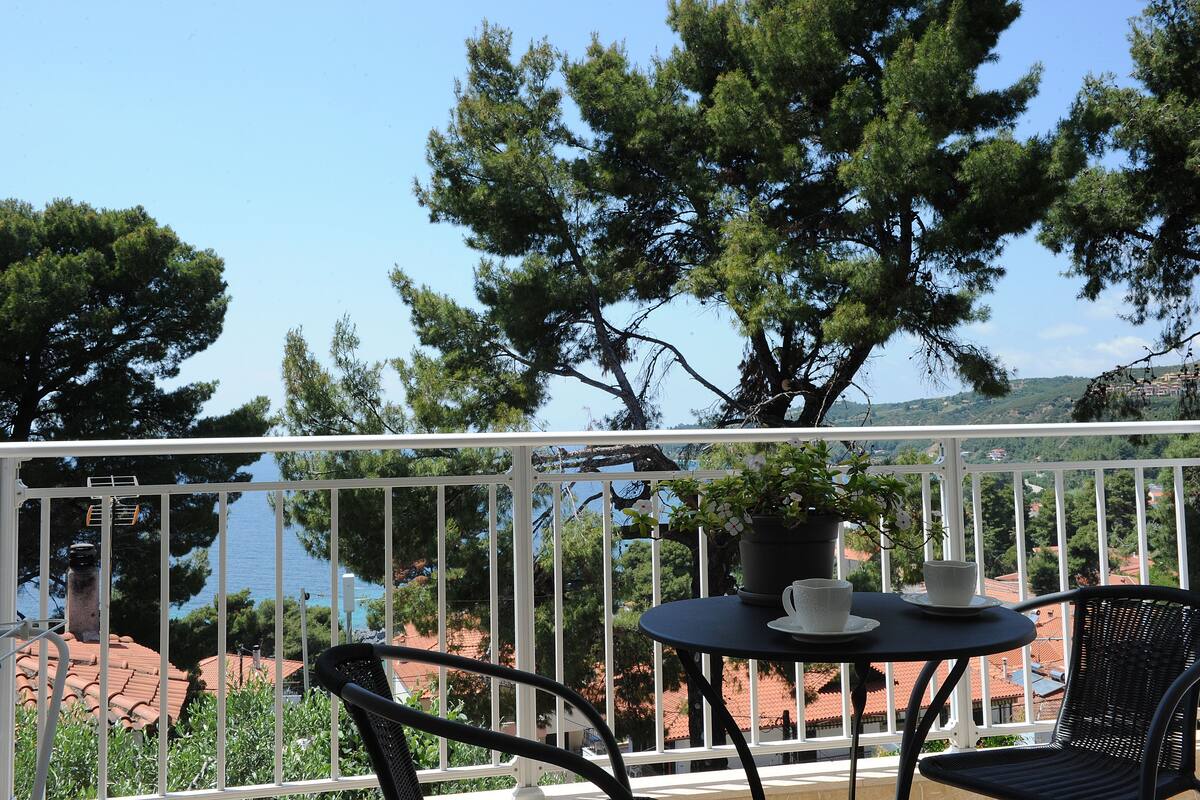A balcony with a table and chairs, overlooking a coastal area with trees and buildings.