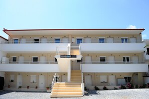 A multi-story building with a sign reading "RODON LOUTRA Apartments." It features a central staircase, multiple balconies, and a series of doors and windows on each floor. - Rodon Loutra (Kassandra)