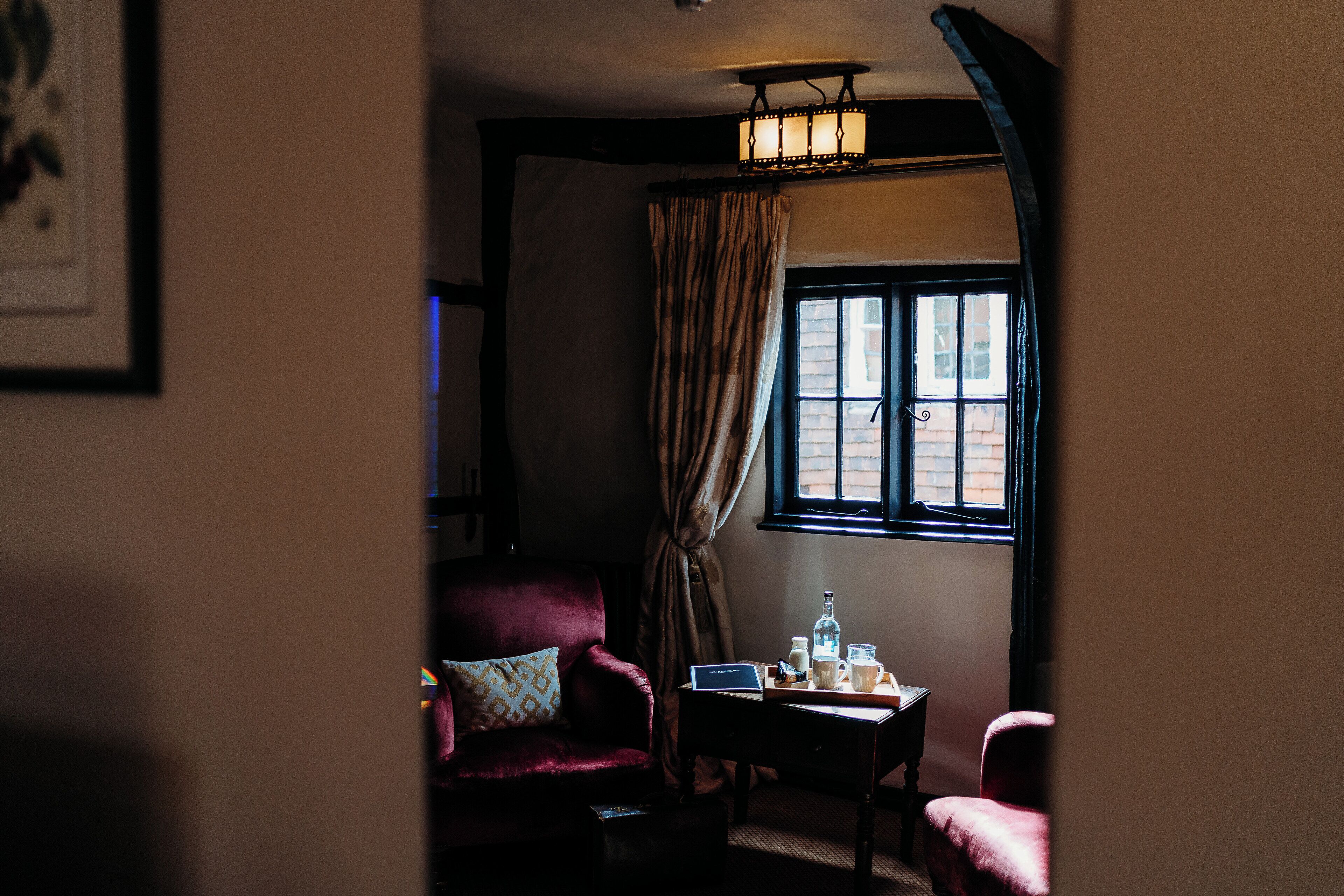 A dimly lit room with a red armchair, a small table with a bottle and glasses, and a window with curtains.