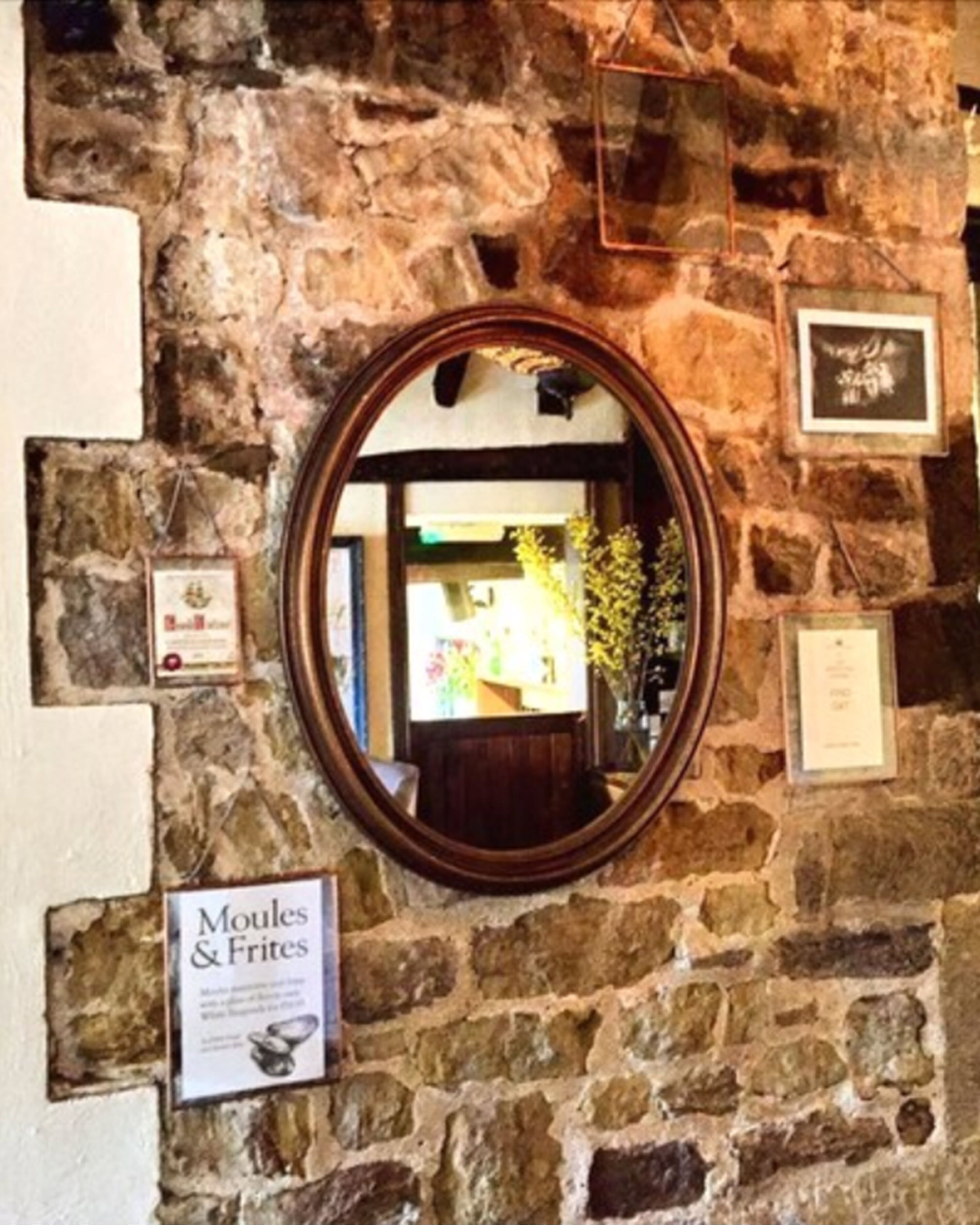 Pared de piedra con un espejo que refleja el interior de una habitación, un cartel que dice 'Moules & Frites' y cuadros enmarcados.