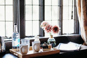 A wooden tray with a bottle, glasses, a mug, and a small box of cookies on a table by a window with a view of a building.