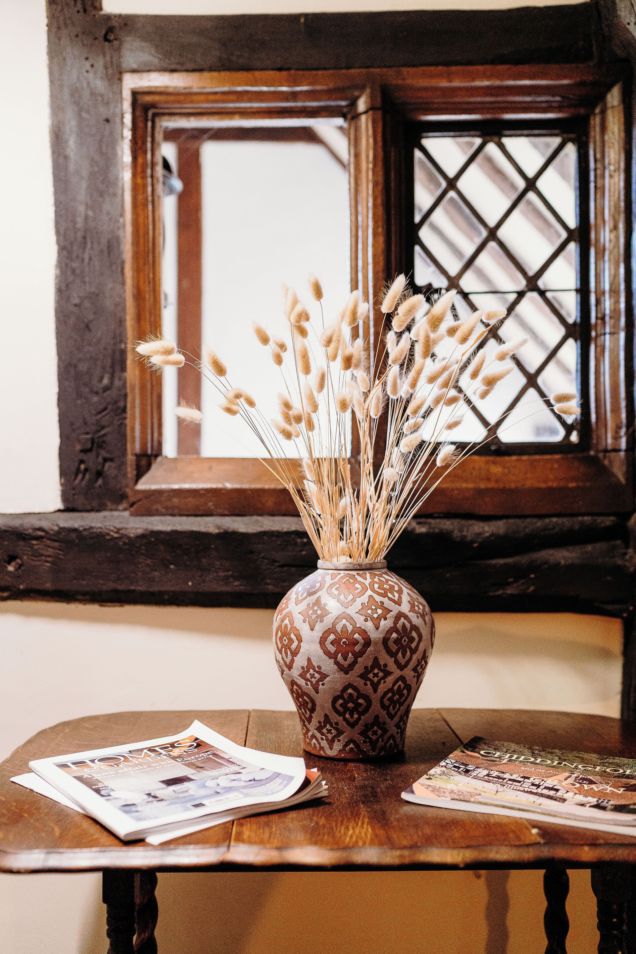 A wooden table with a patterned vase containing dried plants, and two magazines on it.
