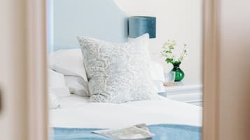 A neatly made bed with white linens and a blue throw blanket, a green vase with flowers on the nightstand, and a white wall with a blue accent.