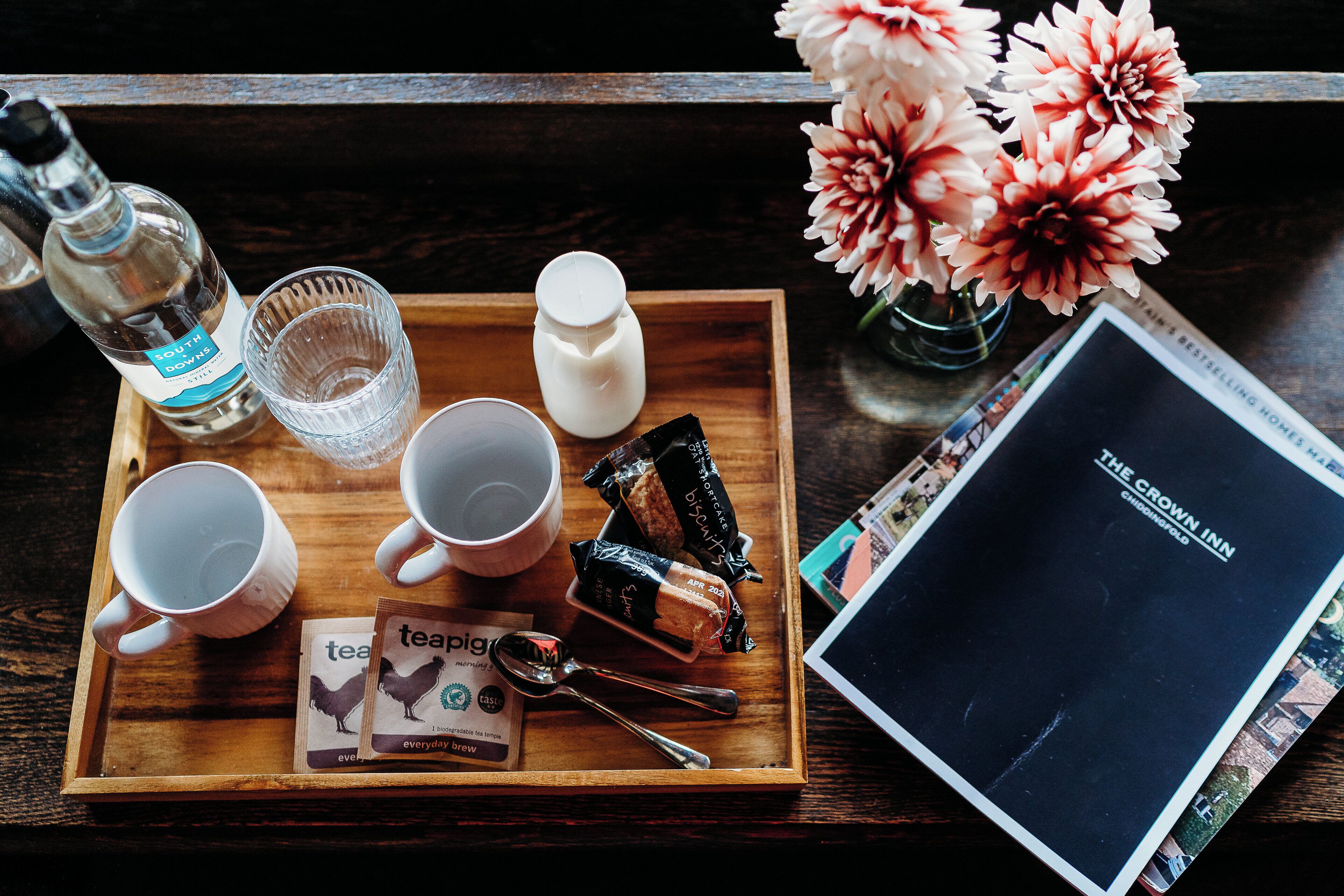 A wooden tray with a bottle of South Downs water, two white cups, a white mug, a small white container, a packet of biscuits, a spoon, and two tea bags. There is also a book titled "The Crown Inn" and a vase with pink flowers.