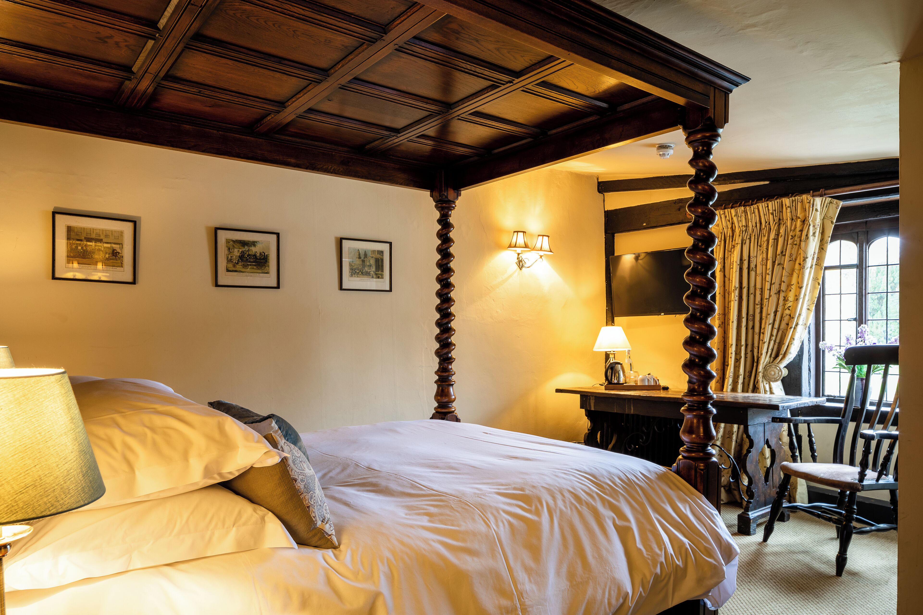 A bedroom with a large bed, wooden ceiling beams, a desk with a lamp, and a chair.