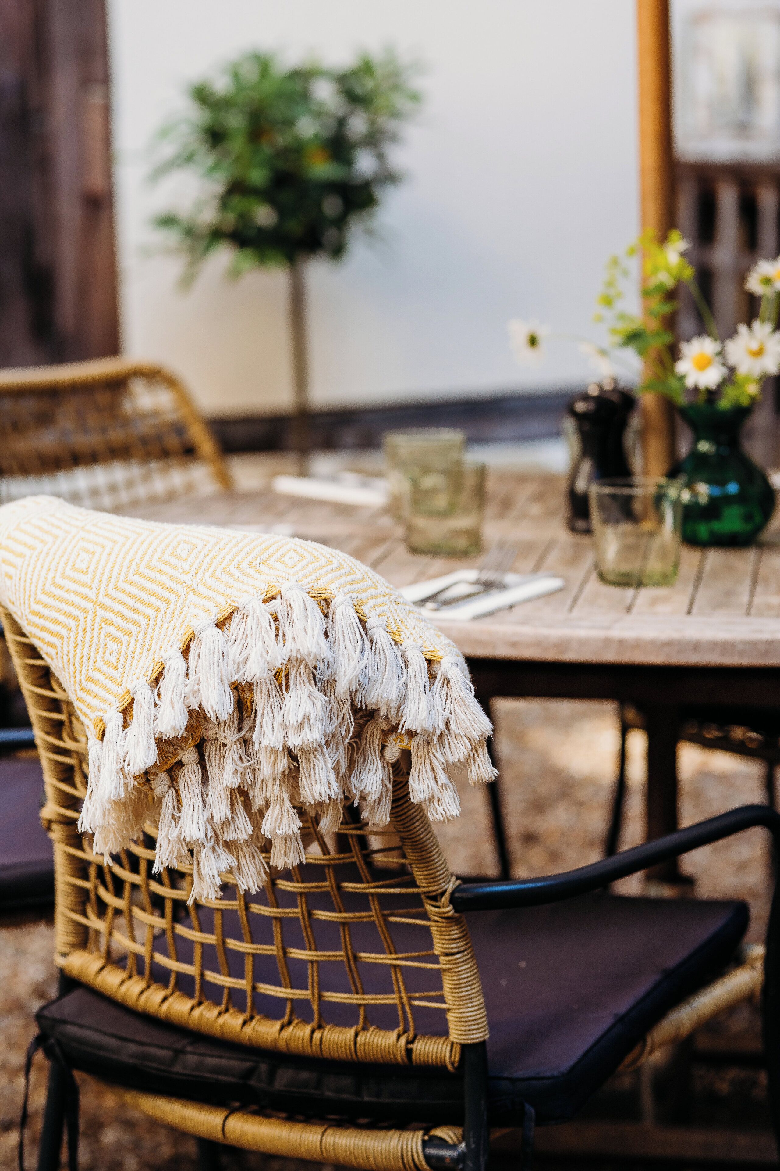 Una silla de mimbre con un plaid con flecos, una mesa de comedor con un florero y una planta en maceta al fondo.