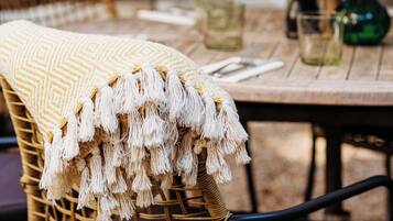 Una silla de mimbre con un plaid con flecos, una mesa de comedor con un florero y una planta en maceta al fondo.