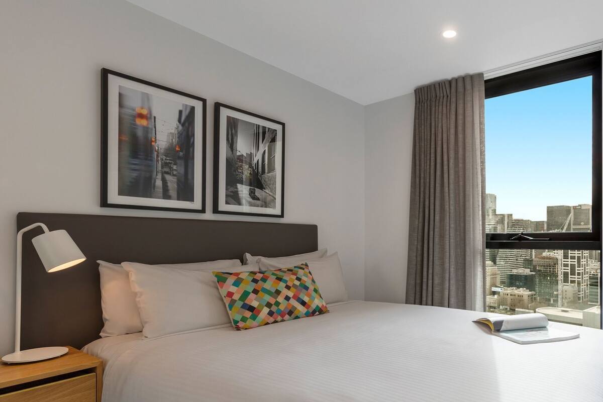 Habitación de hotel con una cama grande, una mesita de noche con lámpara, dos cuadros enmarcados en la pared y vistas de la ciudad a través de una ventana.