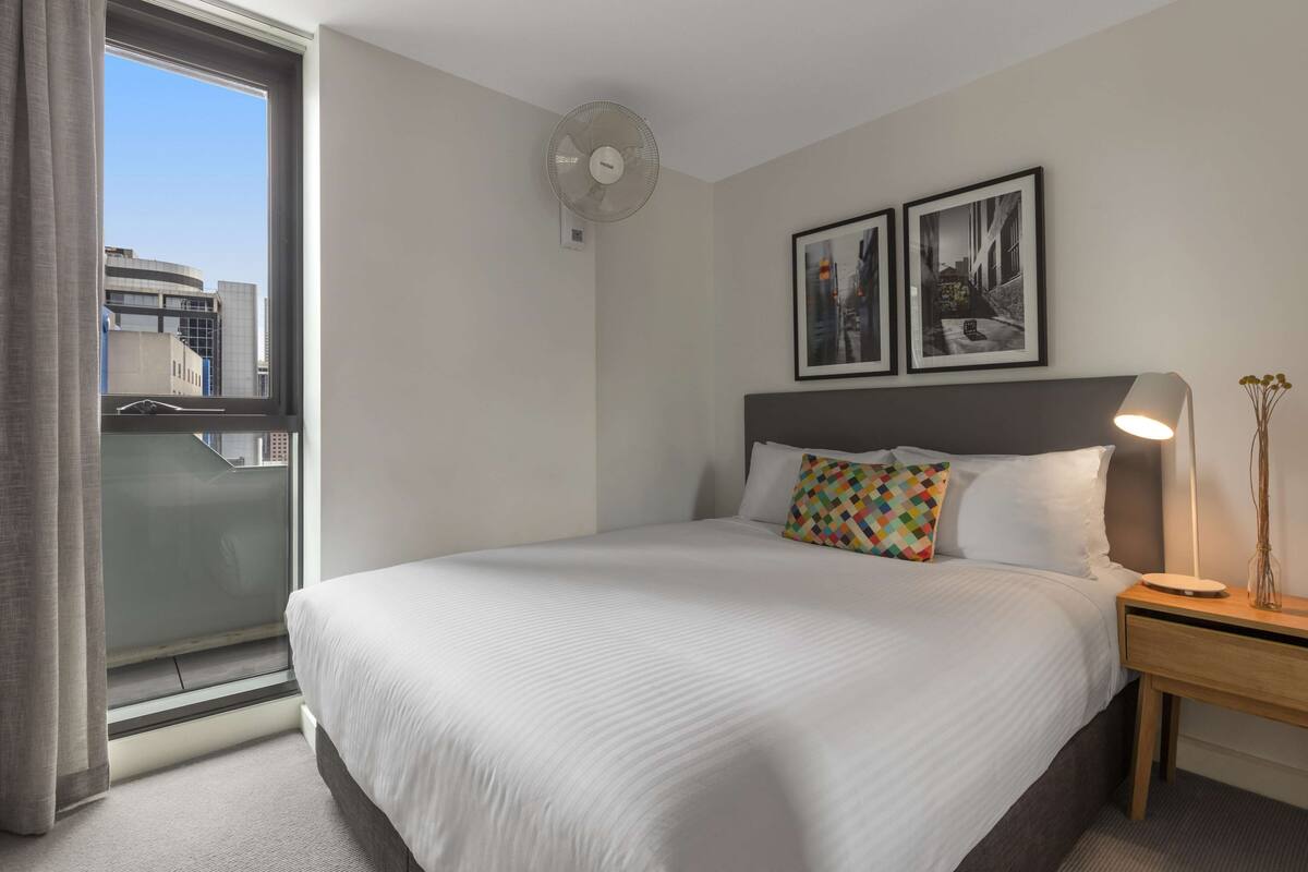 Una habitación de hotel con una cama, una lámpara de noche, un ventilador de pared y dos cuadros enmarcados.