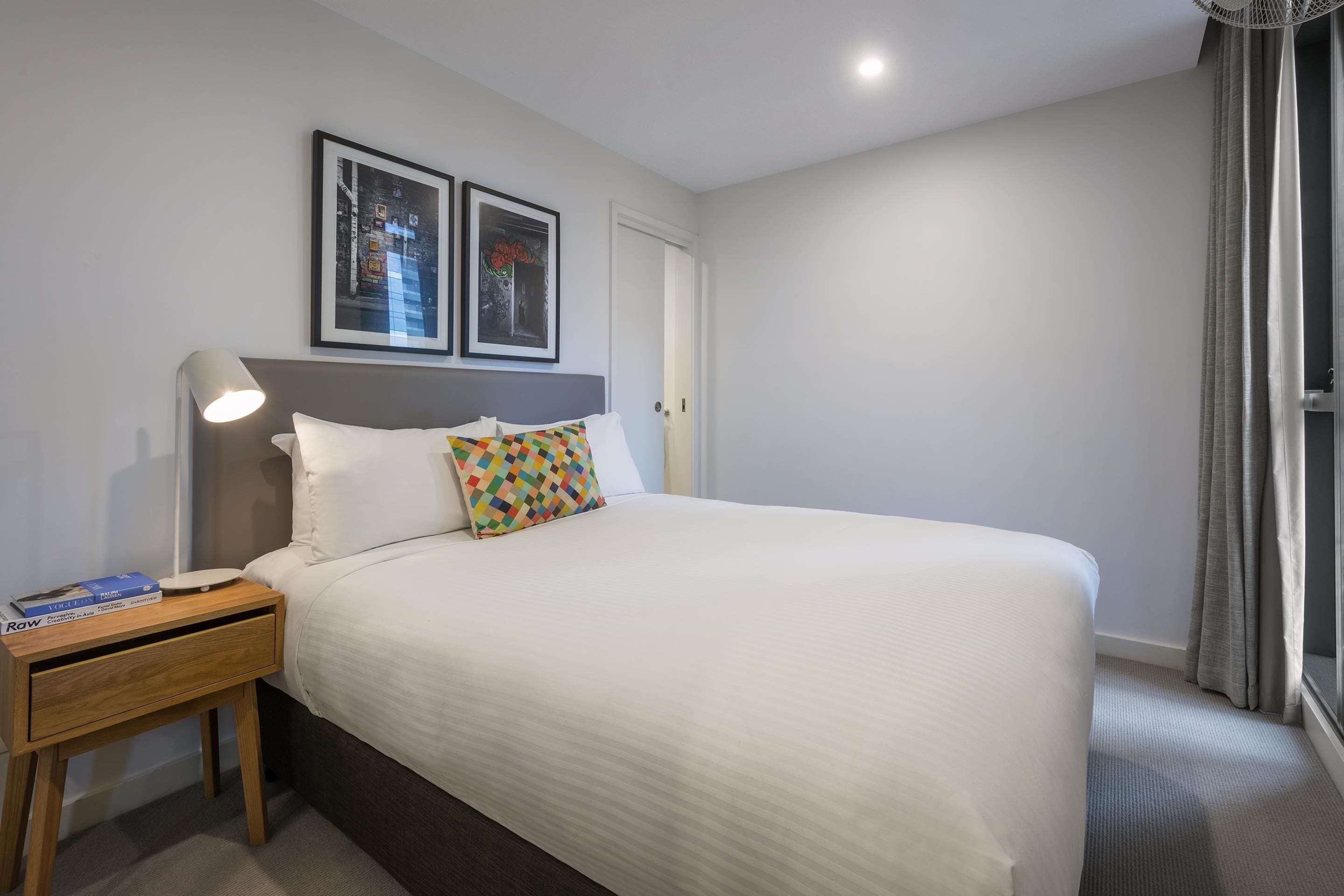 A neatly made bed with a white comforter, a colorful geometric pillow, and a small wooden nightstand with a lamp.