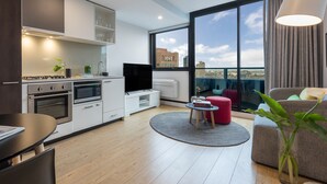 A modern apartment with a kitchen, dining area, and living room. It features a large window with a city view, a flat-screen TV, and a cozy seating area with a red ottoman and a potted plant. - Oaks Melbourne Southbank Suites (Southbank)