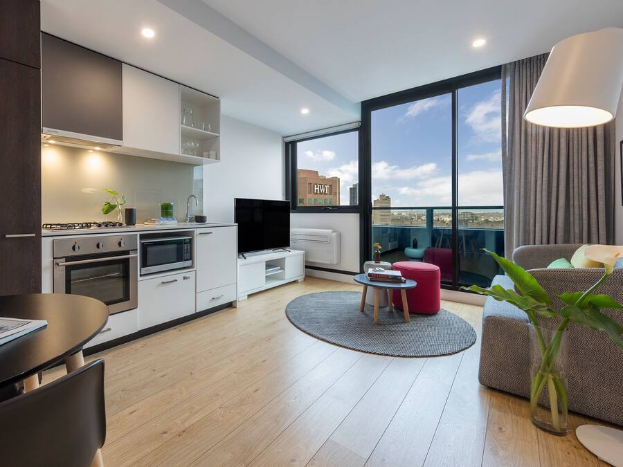 Um apartamento moderno com cozinha, sala de jantar e sala de estar. Possui uma janela grande com vista para a cidade, televisão de tela plana e uma área de estar aconchegante com um banquinho vermelho e um vaso de planta.