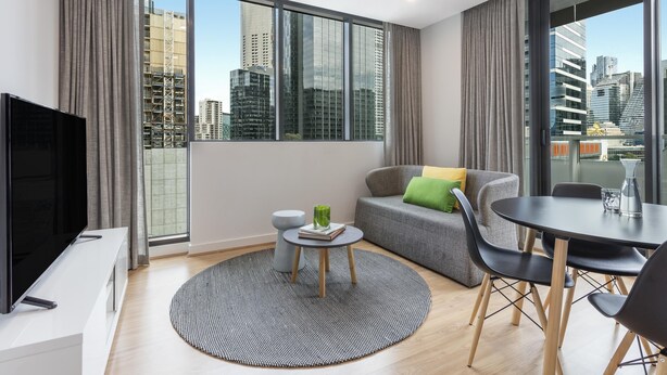 A modern living room with a large window, a flat-screen TV, a round coffee table, a grey sofa, and a dining table with chairs. - Oaks Melbourne Southbank Suites (Southbank)