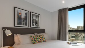 A hotel room with a large bed, a bedside lamp, a nightstand, a window with curtains, and two framed pictures on the wall. - Oaks Melbourne Southbank Suites (Southbank)
