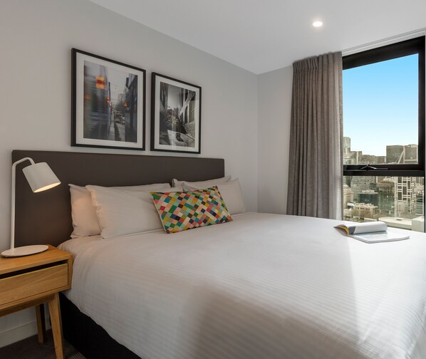 A hotel room with a large bed, a bedside lamp, a nightstand, a window with curtains, and two framed pictures on the wall. - Oaks Melbourne Southbank Suites (Southbank)