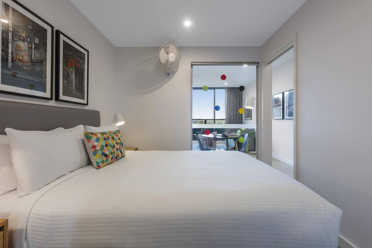 Una cama bien tendida con sábanas blancas y un cojín colorido, un ventilador de pared y cuadros colgados encima.