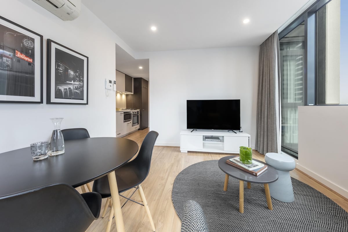 Una sala de estar moderna con un televisor de pantalla plana, una mesa de comedor con sillas, una pequeña mesa redonda con un libro y un vaso, y un amplio ventanal con cortinas.
