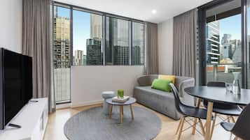 A modern living room with a large window, a flat-screen TV, a round coffee table, a grey sofa, and a dining table with chairs.