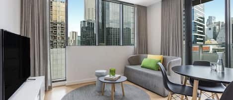 A modern living room with a large window, a flat-screen TV, a round coffee table, a grey sofa, and a dining table with chairs.