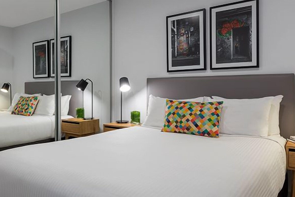 Ein Hotelzimmer mit einem großen Bett, zwei Nachttischen, einem Spiegel und zwei gerahmten Bildern an der Wand.