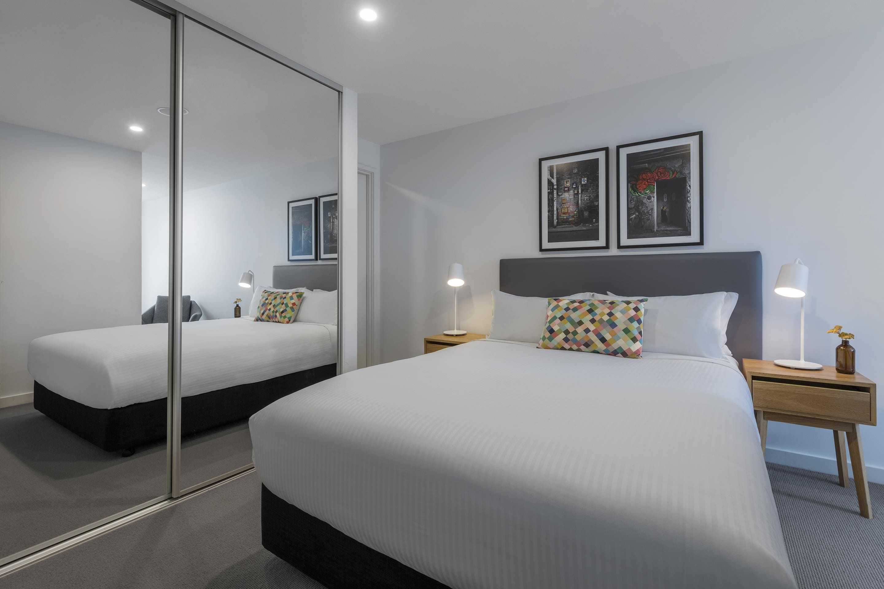 A hotel room with a large bed, a bedside table with a lamp, a mirror, and two framed pictures on the wall.