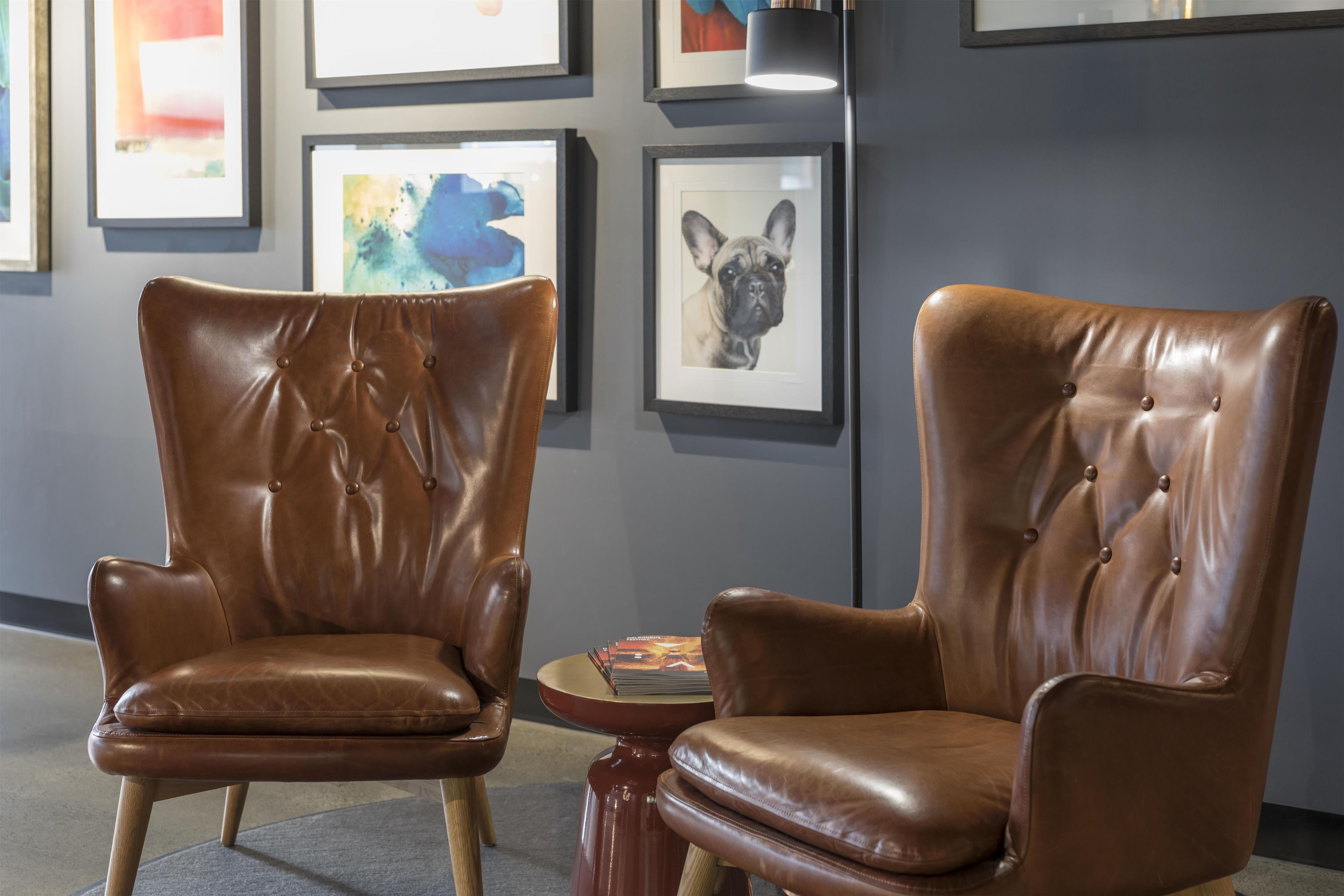 Two brown leather chairs with tufted backs, a small round table with magazines, and framed artwork on the wall.