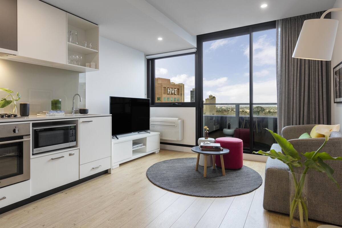 Una sala de estar moderna con televisor de pantalla plana, espacio de cocina y un ventanal con vista a la ciudad.